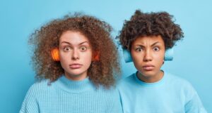 two women, light blue clothing and background, wearing headphones, looking surprised.
