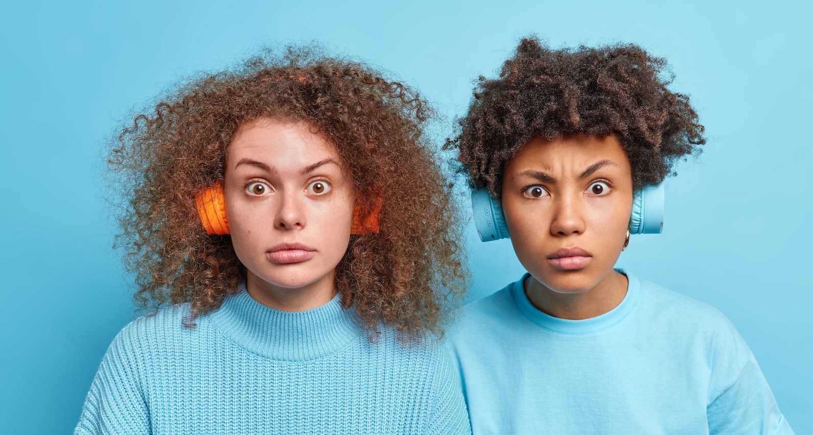 two women, light blue clothing and background, wearing headphones, looking surprised.